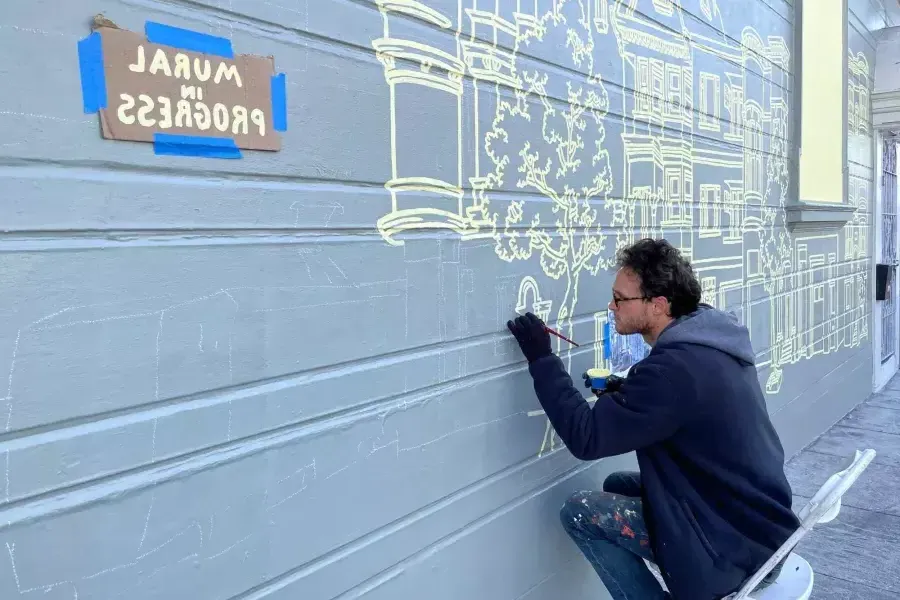 Ein Künstler malt ein Wandgemälde an der Seite eines Gebäudes im Mission District, an dem ein Schild mit der Aufschrift „Mural in Progress“ angebracht ist. 加州贝博体彩app.